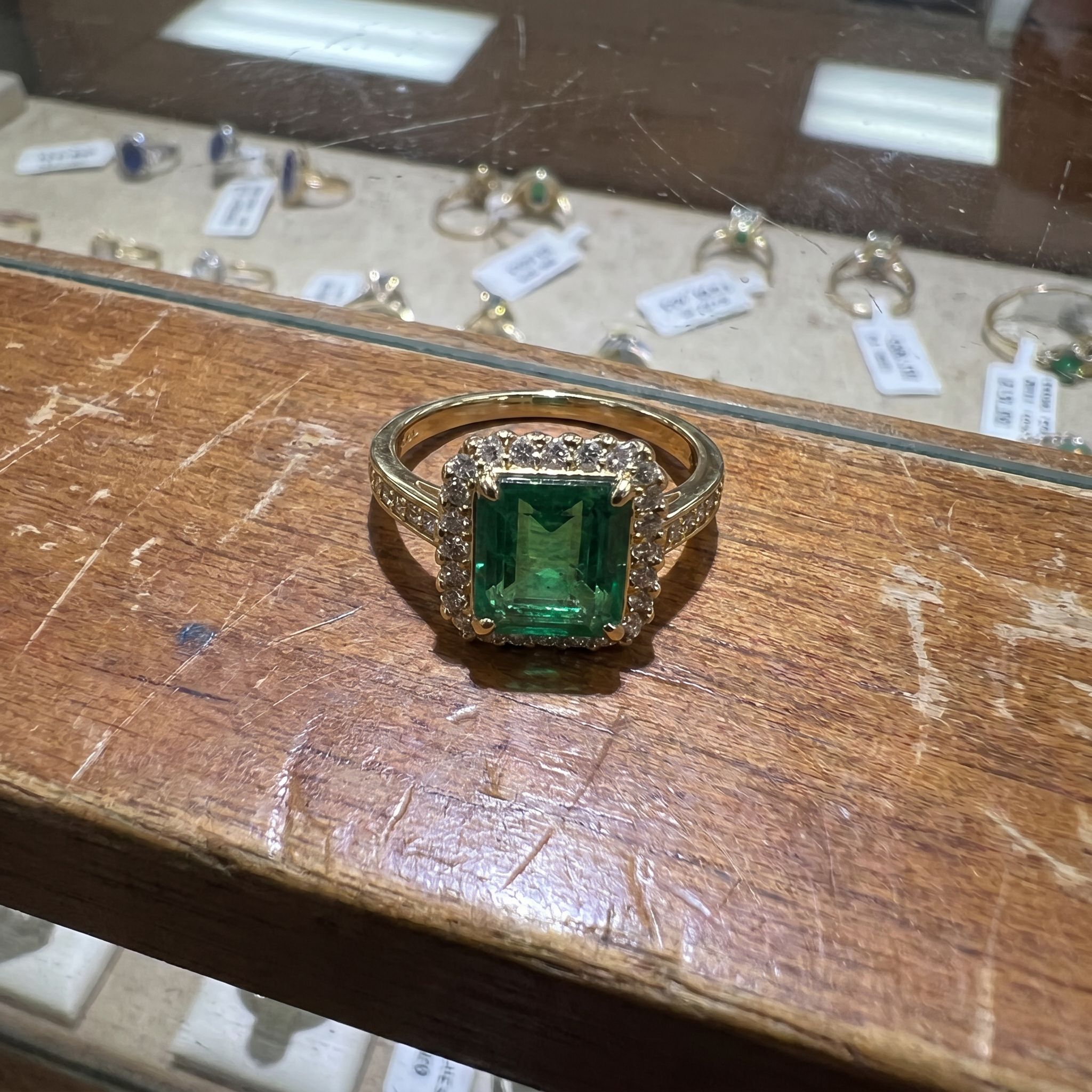 Raw phone snapshot of a gold and emerald halo ring sitting on a wooden jewelry showcase tray.